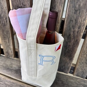 May include: A cream-colored canvas wine tote bag featuring a single handle and an embroidered blue letter R on the front. The bag contains a bottle of wine with a dark red label and a folded pink and white checkered cloth napkin tucked inside.