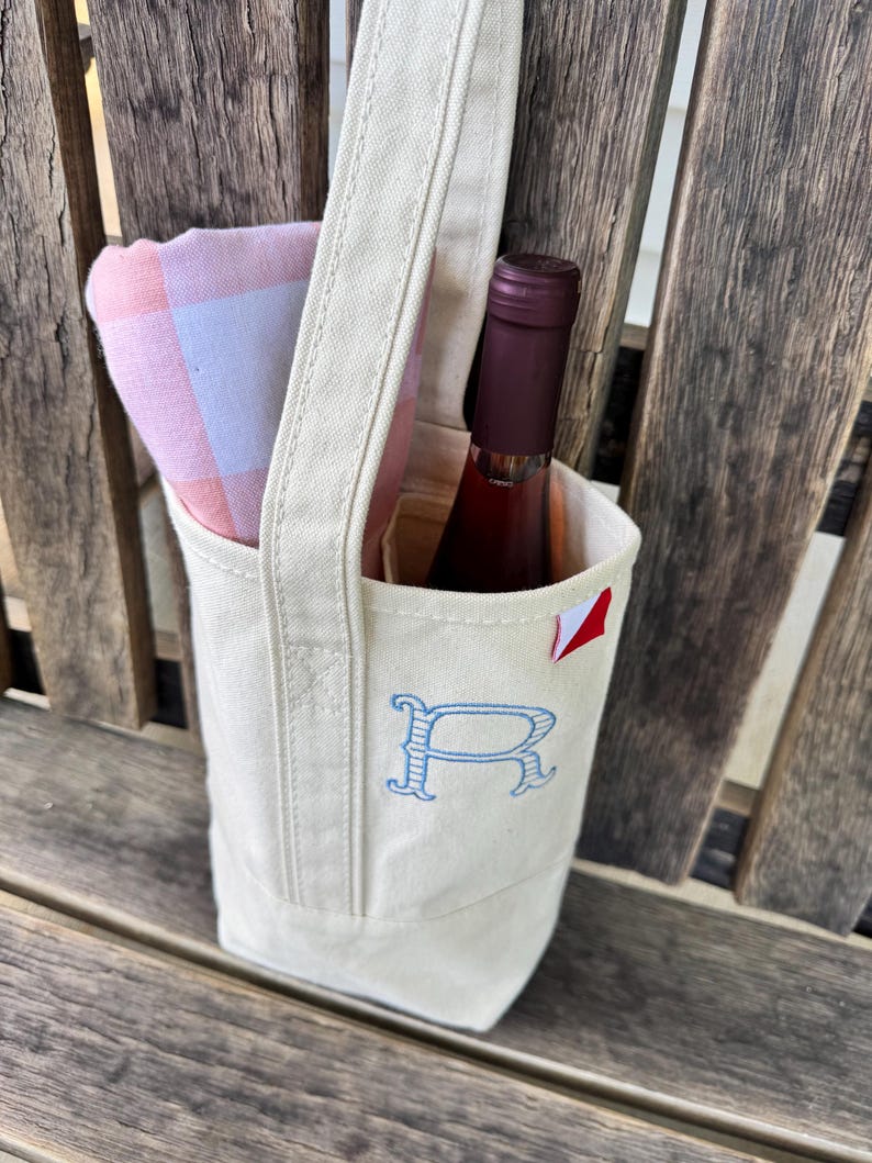 May include: A cream-colored canvas wine tote bag featuring a single handle and an embroidered blue letter R on the front. The bag contains a bottle of wine with a dark red label and a folded pink and white checkered cloth napkin tucked inside.