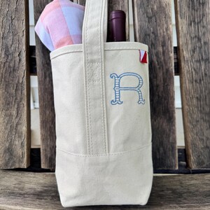 May include: A cream-colored canvas wine tote bag featuring a single vertical handle and a blue embroidered letter R on the front. A dark wine bottle and a folded pink and white checkered cloth are tucked inside the open top of the bag.