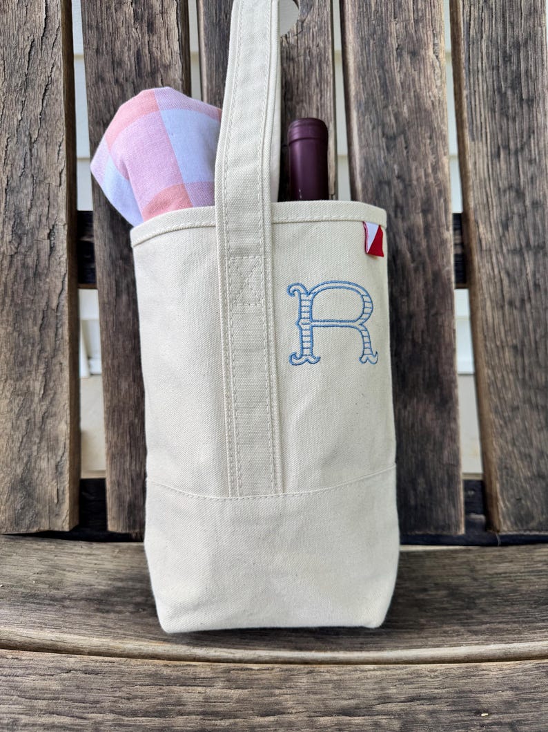 May include: A cream-colored canvas wine tote bag featuring a single vertical handle and a blue embroidered letter R on the front. A dark wine bottle and a folded pink and white checkered cloth are tucked inside the open top of the bag.