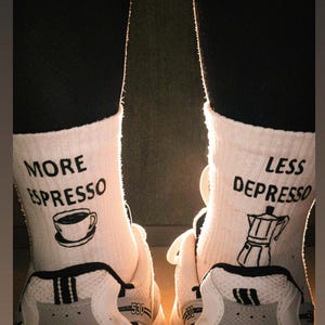 Könnte beinhalten: Ein Paar weiße Socken mit schwarzem Text und Grafiken. Die linke Socke zeigt den Text MORE ESPRESSO mit einer Kaffeetasse, die rechte Socke zeigt LESS DEPRESSO mit einer Espressokanne, getragen in Kombination mit weißen Sportschuhen.
