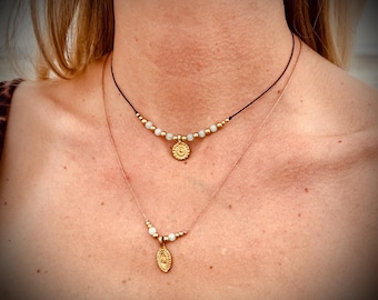 May include: Two layered necklaces with gold pendants and small white beads. The top necklace has a round pendant, while the bottom necklace has an oval pendant. Both necklaces are on a person's neck.