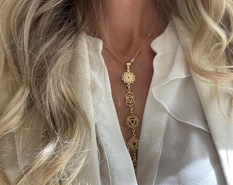 May include: A gold-colored necklace with a series of chakra symbols hanging vertically. The necklace is worn over a white collared shirt. The symbols are intricate and detailed, with a delicate design.
