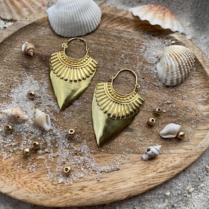 Könnte beinhalten: Ein Paar goldfarbene, schildförmige Ohrringe mit einem Sonnenstrahl-Design oben. Die Ohrringe werden auf einem Holztablett mit Muscheln und kleinen Goldperlen präsentiert, was eine Strandästhetik erzeugt.