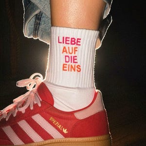 May include: A white ribbed crew sock featuring the red text "LIEBE AUF DIE EINS" worn with a red and light pink trainer that has a gum rubber sole and the word "SPEZIAL" printed in gold on the side, paired with blue denim fabric.