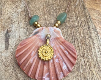 May include: A seashell pendant necklace with a gold-colored charm. The necklace features a natural, peach-colored scallop shell, green beads, and a gold-colored charm. The necklace is strung on a thin, brown cord.