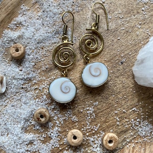 Peut inclure: Boucles d'oreilles en spirale dorées avec des pendentifs en coquillage blancs et beiges. Les boucles d'oreilles sont présentées sur une surface en bois avec du sable et de petites perles en bois. Fermeture à crochet.
