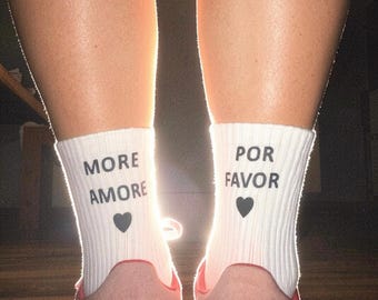 May include: A pair of white ribbed crew socks featuring black text and a small heart icon. The left sock displays the words MORE AMORE above a heart, while the right sock displays the words POR FAVOR above a heart. The socks are worn with pink sneakers.
