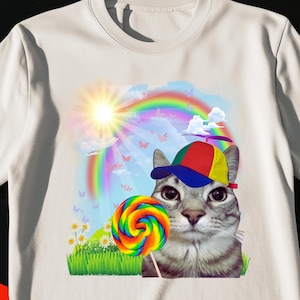 Könnte beinhalten: Ein weißes T-Shirt mit kurzen Ärmeln und einem Aufdruck einer grauen getigerten Katze, die eine bunte Propellermütze trägt. Die Katze hält einen regenbogenfarbenen Lutscher auf einer grünen Wiese unter einer Sonne, einem Regenbogen und Schmetterlingen.