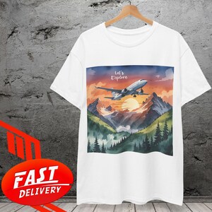 以下が含まれることがあります： 夕焼けの山々の上を離陸する飛行機の水彩画デザインが施された白いTシャツ。「Let's Explore」の文字が飛行機の上にあります。Tシャツは木製のハンガーに掛けられています。