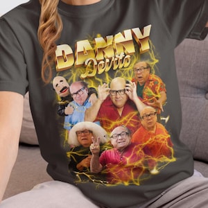 Può includere: T-shirt grigio scuro con un collage di immagini di Danny DeVito e un design di testo dorato "Danny DeVito". Il design include immagini di DeVito in varie pose, con un effetto fulmine giallo.