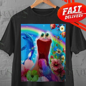 Op de afbeelding: Zwart t-shirt met een grillige afbeelding. Het ontwerp bevat een cartoonachtig wezen met grote ogen, een regenboog, dolfijnen, bloemen en luchtballonnen. Het t-shirt hangt aan een houten hanger.