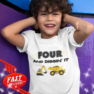 Può includere: T-shirt bianca con la scritta "FOUR AND DIGGIN' IT" in nero e giallo. La maglietta presenta illustrazioni di un escavatore giallo e di un dumper. La maglietta è indossata da un bambino.