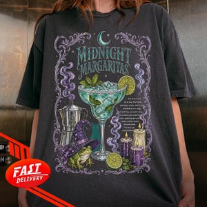 May include: A charcoal gray t-shirt featuring a graphic print with the text Midnight Margaritas. The design includes a margarita glass, a frog wearing a purple witch hat, a silver coffee pot, candles, and smoke swirls in shades of purple and green.