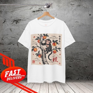 Può includere: T-shirt bianca con grafica di una scimmia su un albero con pesche. Il design include testo giapponese e un timbro rosso. La maglietta è appesa a una gruccia di legno.