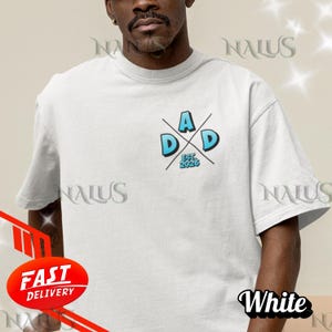 Può includere: T-shirt bianco con grafica blu e bianca con la scritta "DAD EST. 2026". Il testo è centrato sul petto. La maglietta è a girocollo e maniche corte.