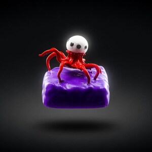 May include: A 3D rendering of a small, red octopus-like creature with a white head and black eyes, perched atop a textured, purple cube. The creature has multiple tentacles and is set against a dark gray background.