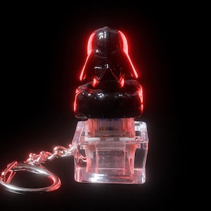 May include: A black Darth Vader keychain with a red glow. The keychain features a small, detailed bust of Darth Vader atop a clear, rectangular base. A silver chain and ring are attached for easy carrying. The background is solid black.