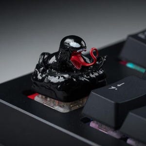 May include: A black Venom-themed keycap with a red tongue and white teeth, set on a black keyboard. The keycap is a detailed, sculpted figure of the Marvel Comics character. The keyboard keys are black.
