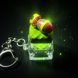 May include: A novelty keychain featuring a miniature bomb resting on a green, textured base within a clear, rectangular case. The bomb is painted with red, yellow, and brown details. A silver-colored chain and clasp are attached.