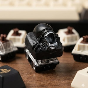 Może przedstawiać: Czarny keycap z motywem Obcego z szczegółowym, otwartym projektem ust, ukazującym ostre zęby. Klawisz otoczony jest innymi klawiszami klawiatury z brązowymi i białymi akcentami, umieszczonymi na drewnianej powierzchni. Ten unikalny keycap dodaje charakterystycznego akcentu do każdej klawiatury.
