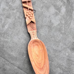 May include: A wooden spoon with a carved design of grapes and leaves. The spoon is brown and has a long handle.