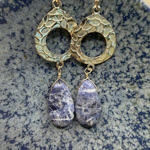 May include: A pair of gold-colored earrings with a teardrop shape. The top portion has a textured, open design. The bottom features a blue and white stone in a teardrop shape, suspended by a gold wire.