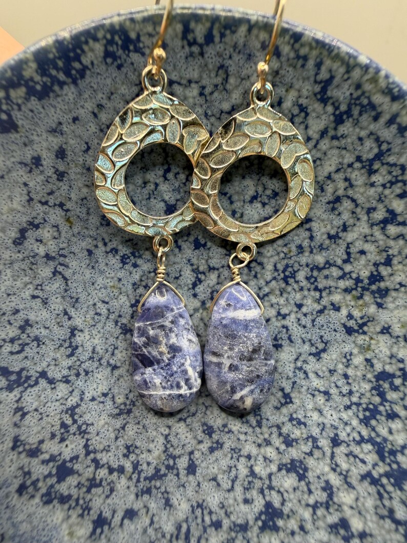 May include: A pair of gold-colored earrings with a teardrop shape. The top portion has a textured, open design. The bottom features a blue and white stone in a teardrop shape, suspended by a gold wire.