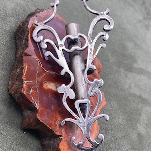 May include: A silver filigree pendant with a decorative design and a small tube in the center. The pendant is on a brown and white stone background.