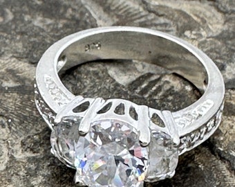 May include: A silver ring with a large, clear, round cubic zirconia stone surrounded by two smaller cubic zirconia stones. The band is set with smaller cubic zirconia stones.