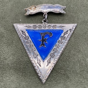 May include: A silver triangular pendant with a blue enamel center featuring a black letter 'F'. The pendant has a silver ribbon attached to the top.
