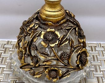 May include: A clear glass perfume bottle with a gold floral design. The bottle has a round body and a faceted base.