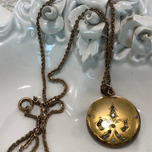 May include: A gold locket necklace with a fleur-de-lis design and small clear stones. The chain is a delicate, linked style.