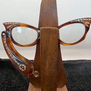 May include: Brown cat-eye glasses with rhinestone embellishments on the frame and arms. The glasses are displayed on a wooden stand, showcasing their vintage style and design.