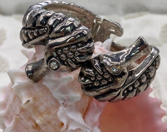 May include: A pair of silver tone hinged bracelets with a detailed design of a stylized horse head with a single rhinestone on each. The bracelets are resting on a pink seashell.