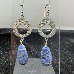 May include: A pair of silver earrings with a textured, open teardrop design. Each earring features a teardrop-shaped, blue stone pendant. The earrings are displayed against a neutral background.