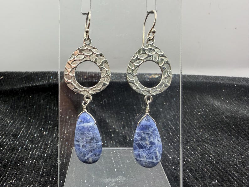 May include: A pair of silver earrings with a textured, open teardrop design. Each earring features a teardrop-shaped, blue stone pendant. The earrings are displayed against a neutral background.