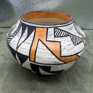 May include: A round ceramic pot featuring a white base with a distressed, crackled texture. The exterior is decorated with bold black geometric shapes, including triangles and lines, alongside prominent orange angular patterns.