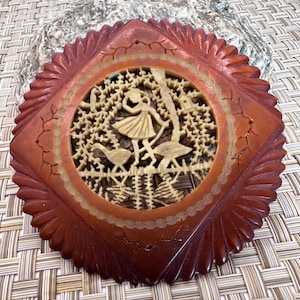 May include: A round, red, carved wooden ornament with a detailed design of a woman in a dress and birds in a tree. The ornament has a scalloped edge.