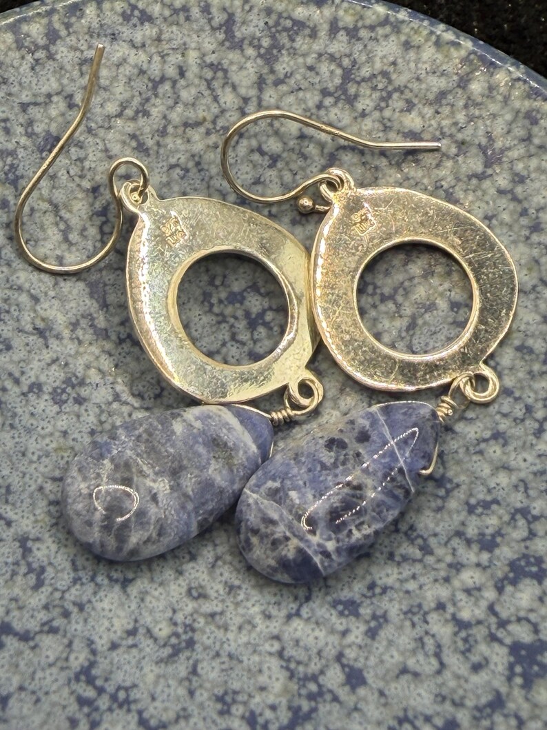 May include: Silver drop earrings with teardrop-shaped blue sodalite stones. The earrings have a circular design with a small loop at the top for the ear wire. The sodalite stones are a deep blue with white marbling.