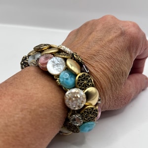 May include: A bracelet made of a variety of buttons in different colors and textures, including gold, white, pink, and blue. The bracelet is worn on a person's wrist.