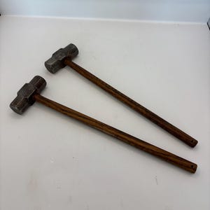 May include: Two sledgehammers with dark wooden handles and gray metal heads are displayed against a white background. The hammers are positioned diagonally, showcasing their construction and design. The tools are likely used for heavy-duty tasks.