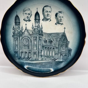 May include: A blue and white ceramic plate with a black rim and gold trim. The plate features a black and white illustration of a church building with three men's portraits above it.