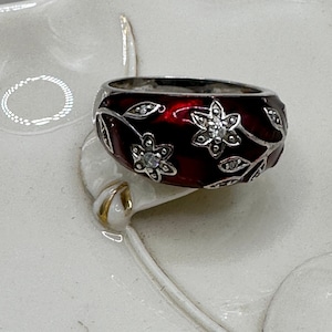 May include: A silver ring with a wide band featuring a deep red enamel design. The ring is adorned with floral and leaf patterns, accented with small, clear gemstones. The flowers and leaves are set against the red enamel, creating a striking contrast.