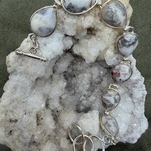 May include: A silver bracelet featuring seven teardrop-shaped stones with swirling white, gray, and brown patterns. The stones are set in silver bezels and connected by silver links, finished with a toggle clasp and three circular adjustment rings.