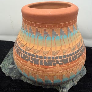 May include: A terracotta-colored ceramic vase with a wide opening and a rounded body. The vase features a carved design with bands of orange, blue, and gray geometric patterns. The vase sits on a gray stone base.