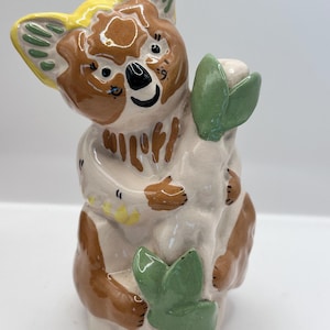 May include: A ceramic koala figurine with brown, yellow, and green accents. The koala is holding a white object with green leaves. The figurine has a smiling face and is painted with a glossy finish.