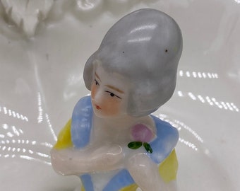 May include: A porcelain figurine of a woman wearing a blue and yellow dress and a gray hat. She is holding a pink flower in her hand. The figurine is sitting on a white porcelain base.
