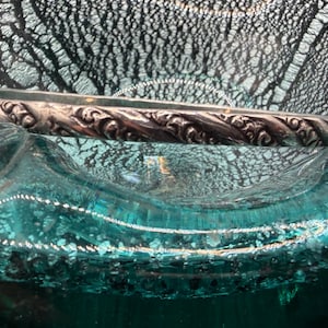 May include: A silver-colored bangle bracelet with an ornate, swirling floral design. The bracelet is resting on a textured, teal-colored glass surface, creating a contrast of textures and colors. The bracelet's intricate details are highlighted against the backdrop.