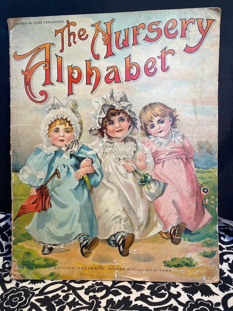 May include: A vintage book cover featuring three children in dresses walking along a path. The book title is "The Nursery Alphabet" printed in red and orange letters. The book cover is printed on linen and is untearable.